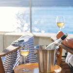 Two deck chairs on a waterfront balcony. A woman is sitting in one of the chairs. There is an ice bucket with wine and glasses. Sun can be seen reflecting on the water.